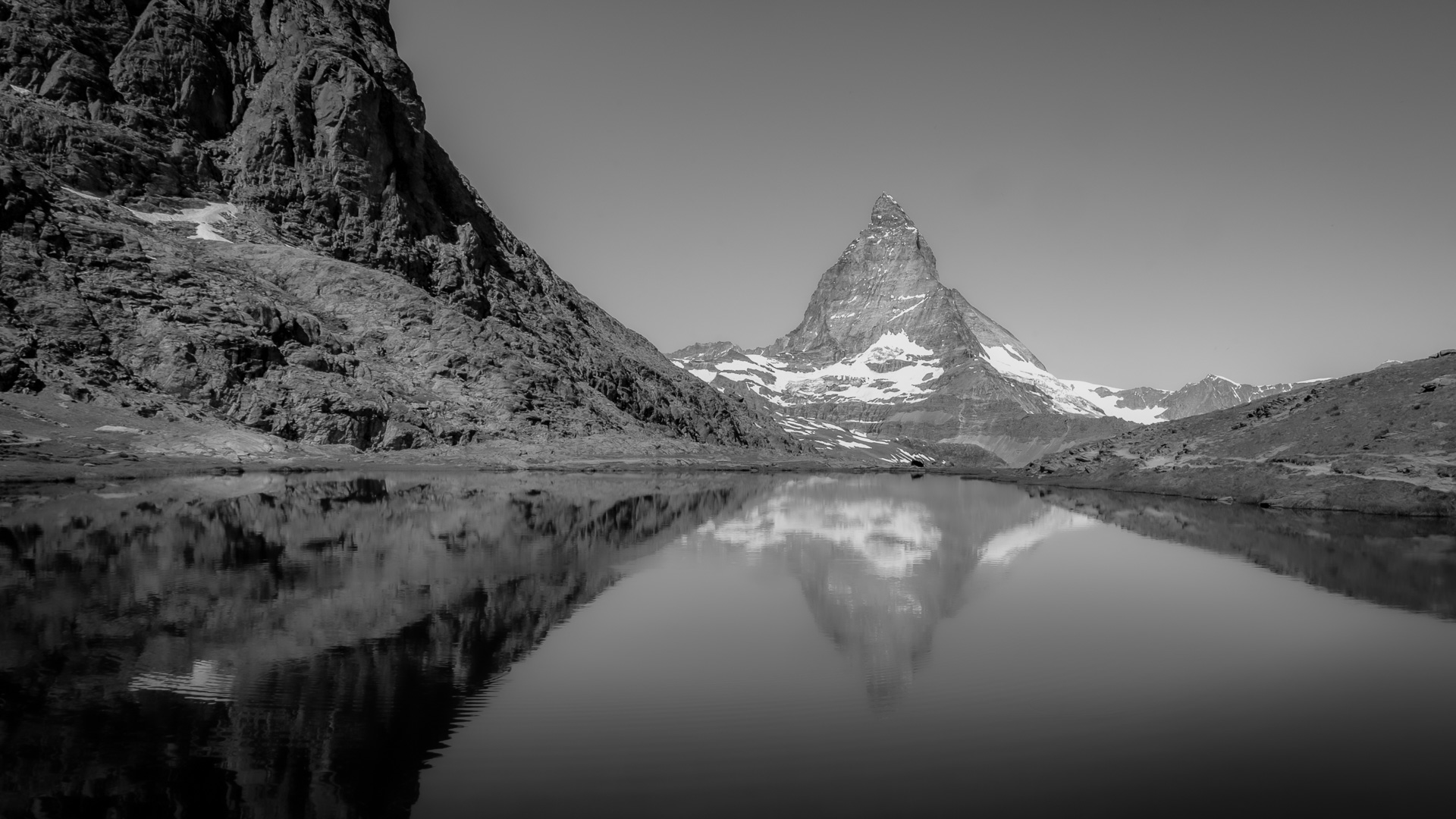 Sort/hvid bjergsø med stille vand, stejle klipper og Matterhorn-lignende top i baggrunden.